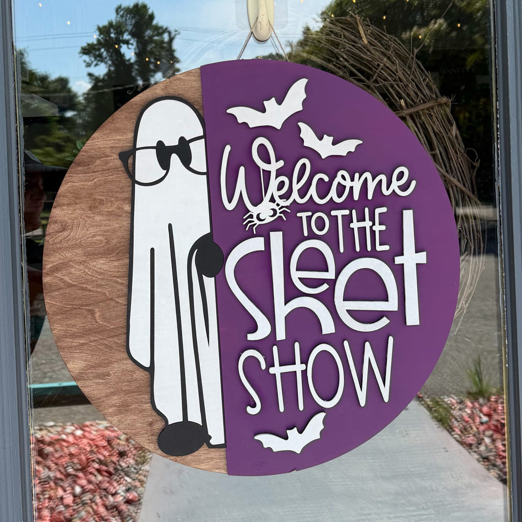 A circular Halloween window decoration hanging on a glass door, featuring a split design with a brown wood-grain side showing a white cartoon ghost and a purple side with white text reading 'Welcome to the Skit Show' and bat silhouettes.