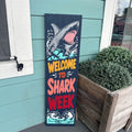 Decorative sign with shark illustration and 'Welcome to Shark Week' text against a teal wall.