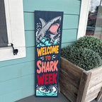 Decorative sign with shark illustration and 'Welcome to Shark Week' text against a teal wall.
