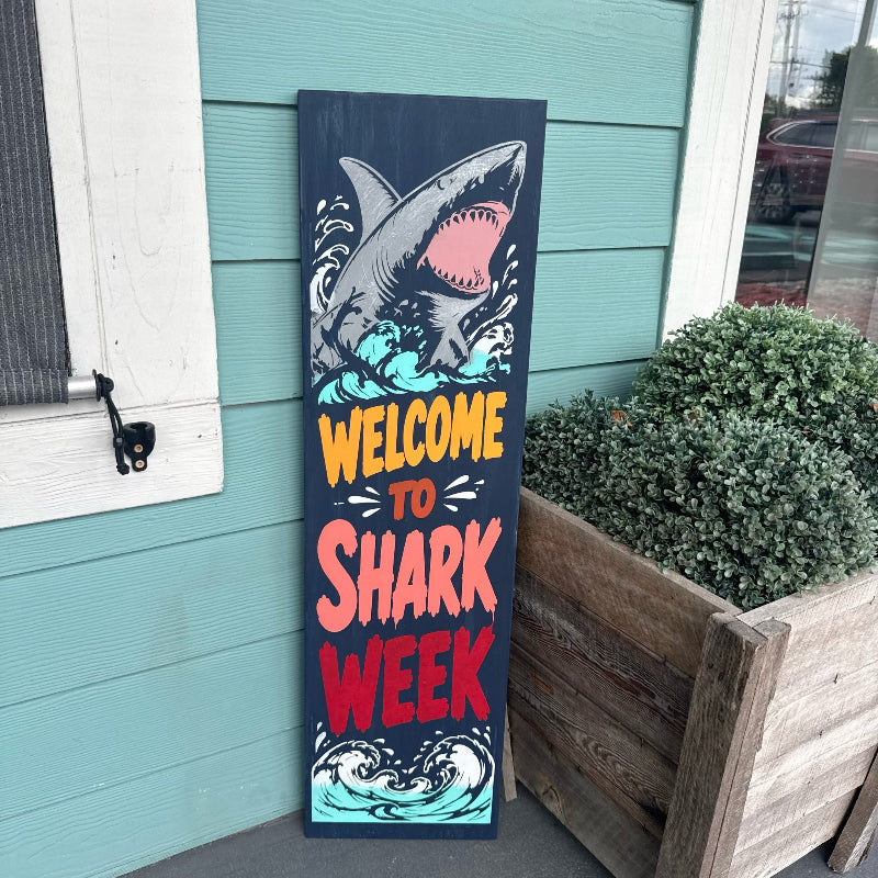 Decorative sign with shark illustration and 'Welcome to Shark Week' text against a teal wall.