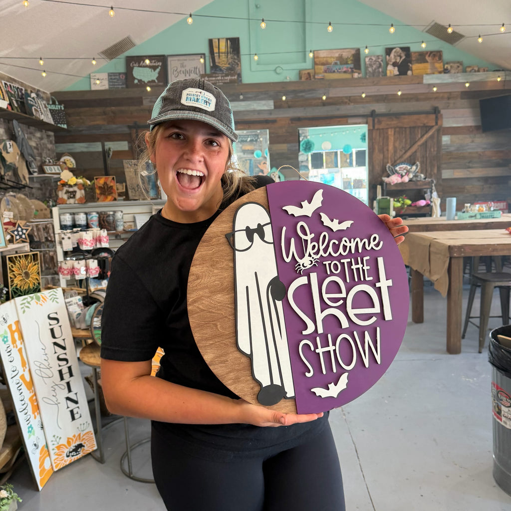 A woman in a black t-shirt and a gray cap holds a round wooden sign that reads 'Welcome to the Sket Show' with a cartoon ghost wearing glasses. The sign is split between a purple section and a natural wood-grain section.