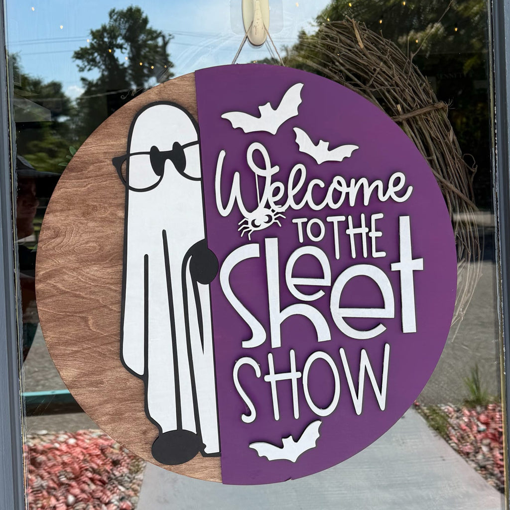 A circular Halloween window decoration hanging on a glass door, featuring a split design with a brown wood-grain side showing a white cartoon ghost and a purple side with white text reading 'Welcome to the Skit Show' and bat silhouettes.