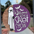 A circular Halloween window decoration hanging on a glass door, featuring a split design with a brown wood-grain side showing a white cartoon ghost and a purple side with white text reading 'Welcome to the Skit Show' and bat silhouettes.