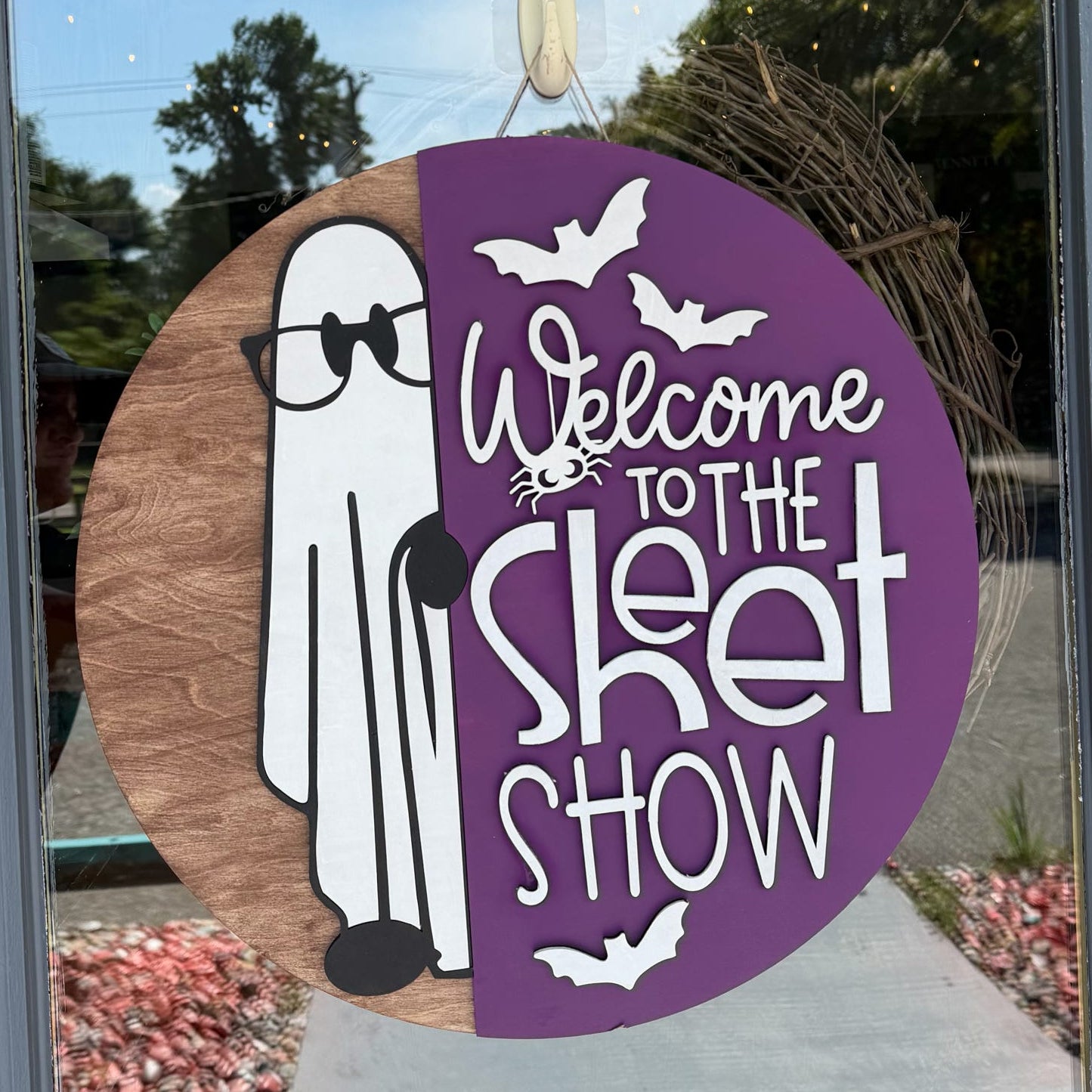 A circular Halloween window decoration hanging on a glass door, featuring a split design with a brown wood-grain side showing a white cartoon ghost and a purple side with white text reading 'Welcome to the Skit Show' and bat silhouettes.