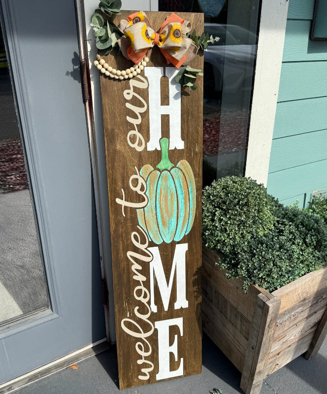 Wooden sign with 'Welcome to our Home' and pumpkin design in front of a door.