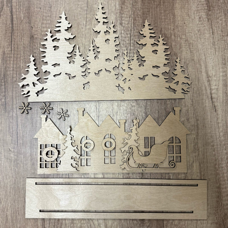 Wooden cutout of a winter scene with houses, trees, and a sleigh on a wooden background