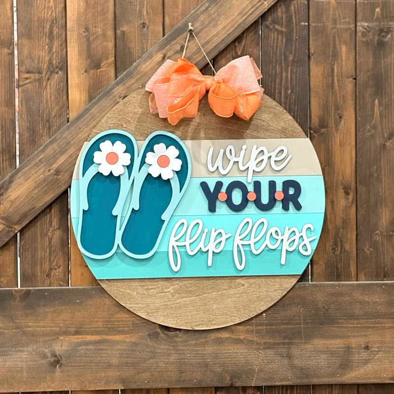 Decorative sign with flip-flop design and text 'wipe YOUR flip flops' on a wooden background