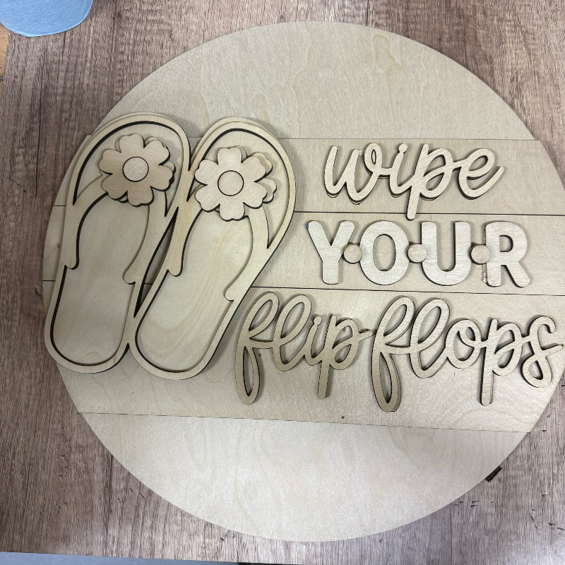 Wooden sign with flip flop design and text 'wipe your flip flops' on a wooden surface