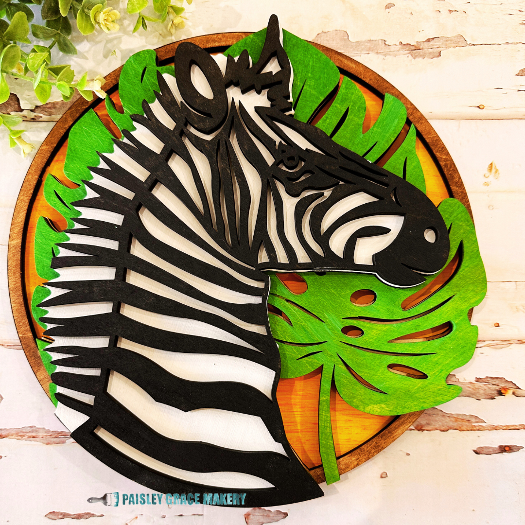 A layered wooden sign in the shape of a zebra's head with black and white stripes, green leaves, and a brown background.