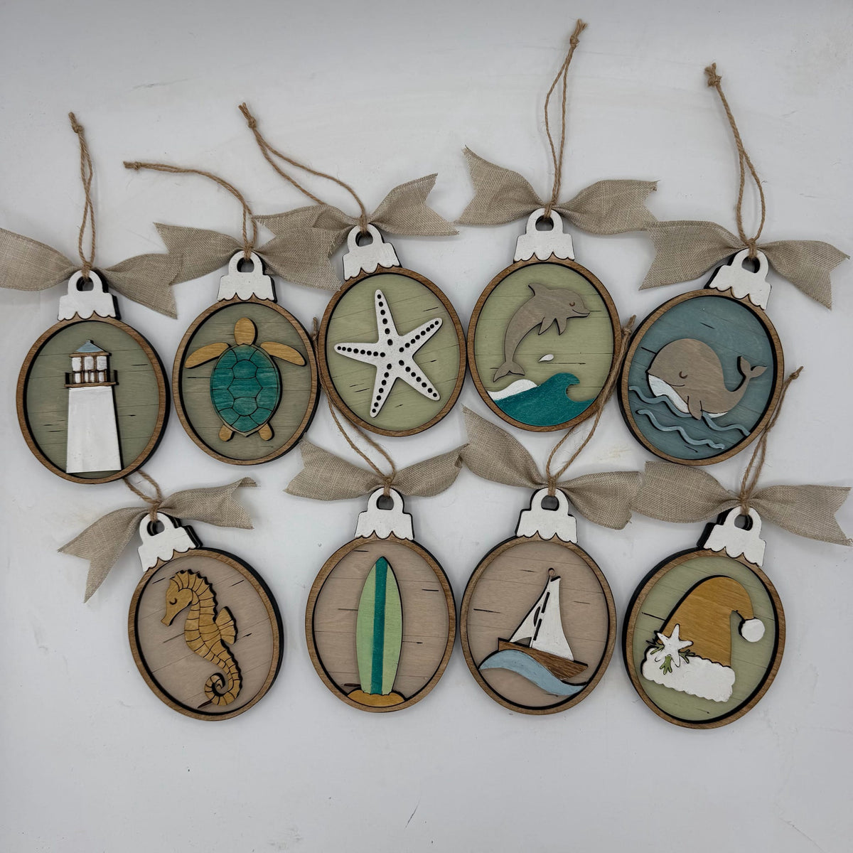A set of nine oval wooden hanging ornaments with burlap ribbon bows, each featuring a different nautical or beach-themed laser-cut design including a lighthouse, turtle, starfish, dolphin, whale, seahorse, surfboard, sailboat, and Santa Claus.