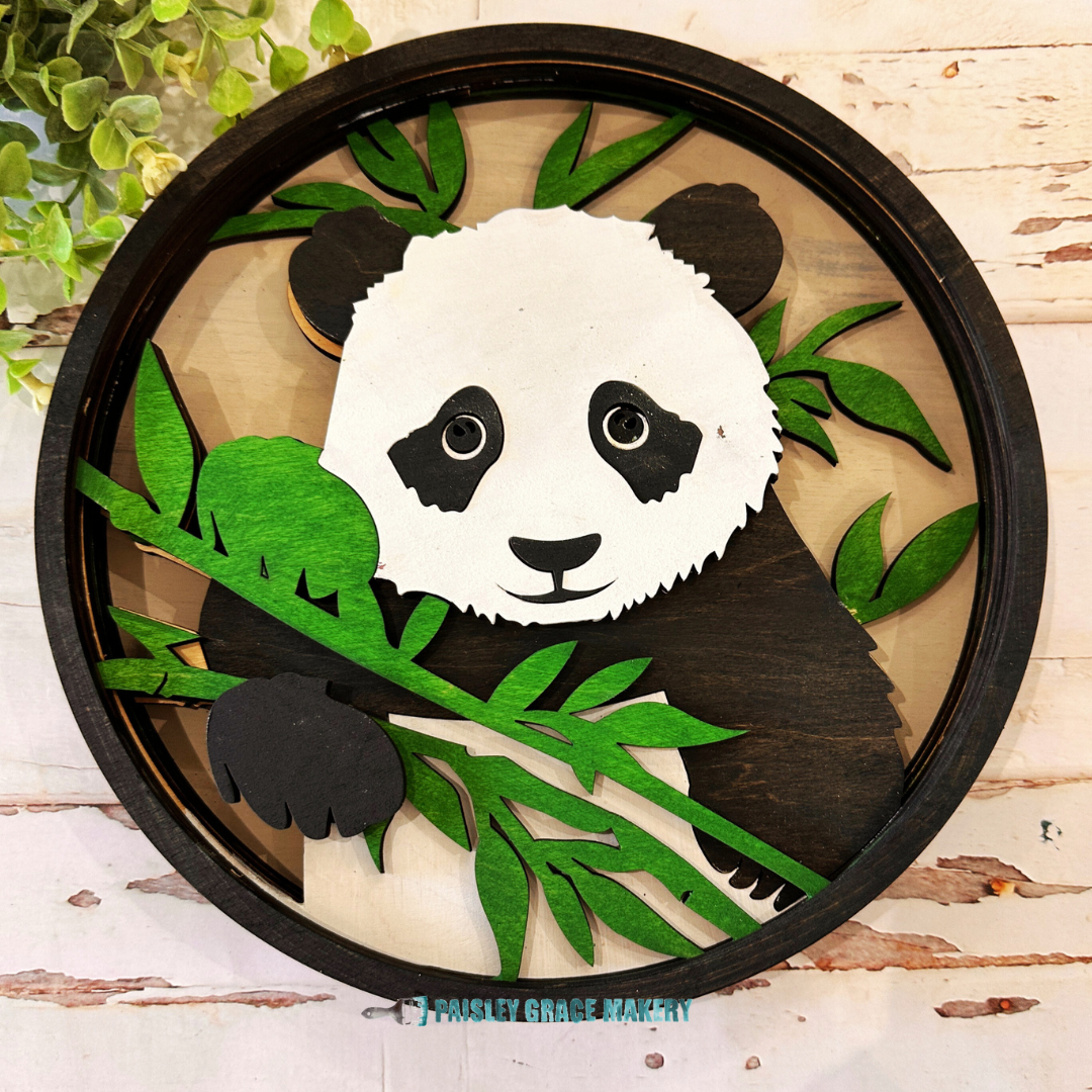 A small round sign featuring a colorful panda layered design with green leaves and a black outline, hanging on a wooden surface.