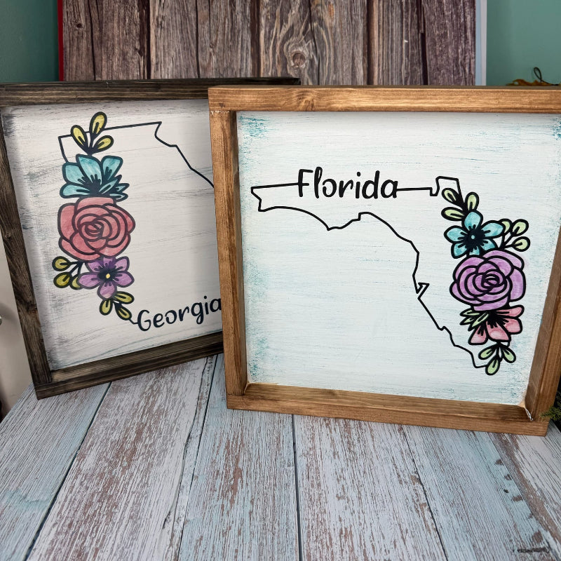 Two framed signs with floral designs and state names on a wooden surface.