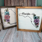 Two framed signs with floral designs and state names on a wooden surface.